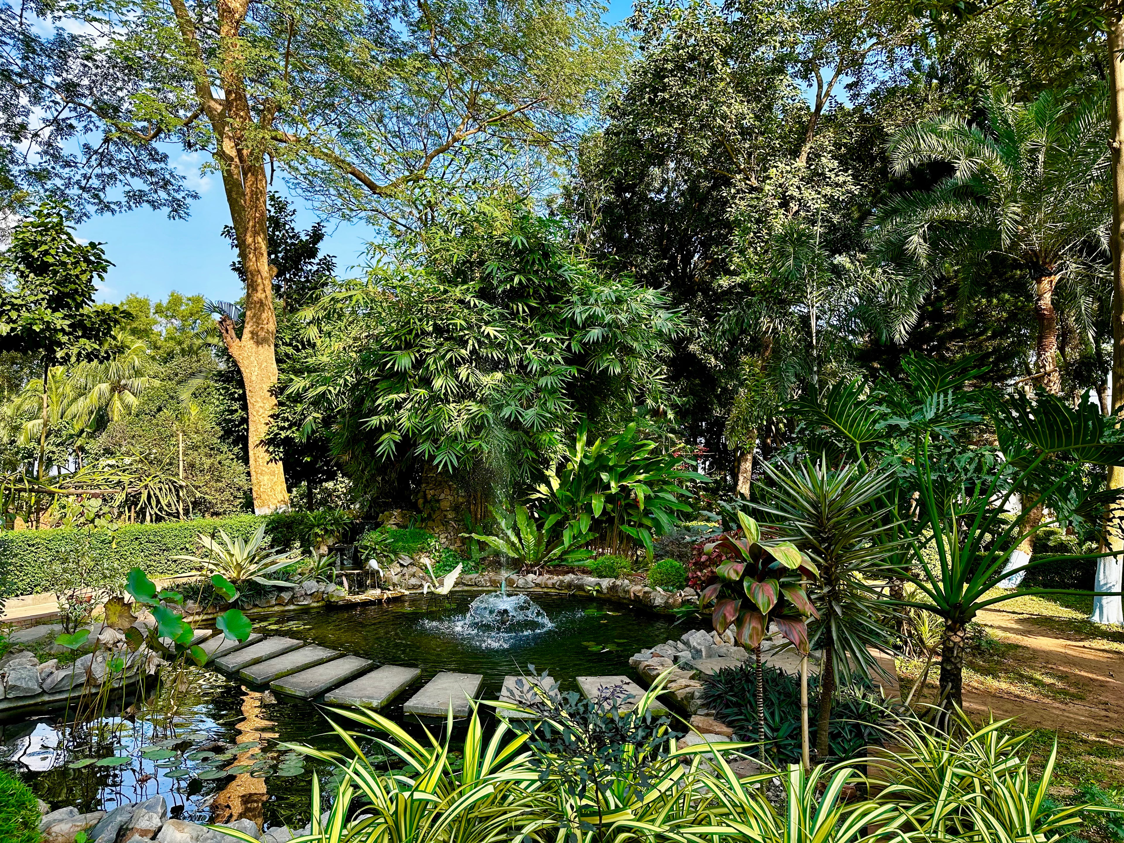 Tranquil pond with stepping stones and a heron statue at Barnochata, an enchanting Dhaka hotel and resort in Savar surrounded by lush foliage.